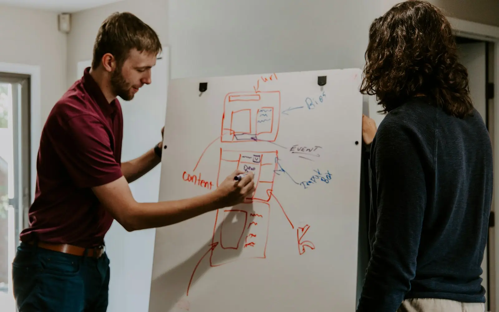 Two people sketching an app screen flow in red marker on a whiteboard